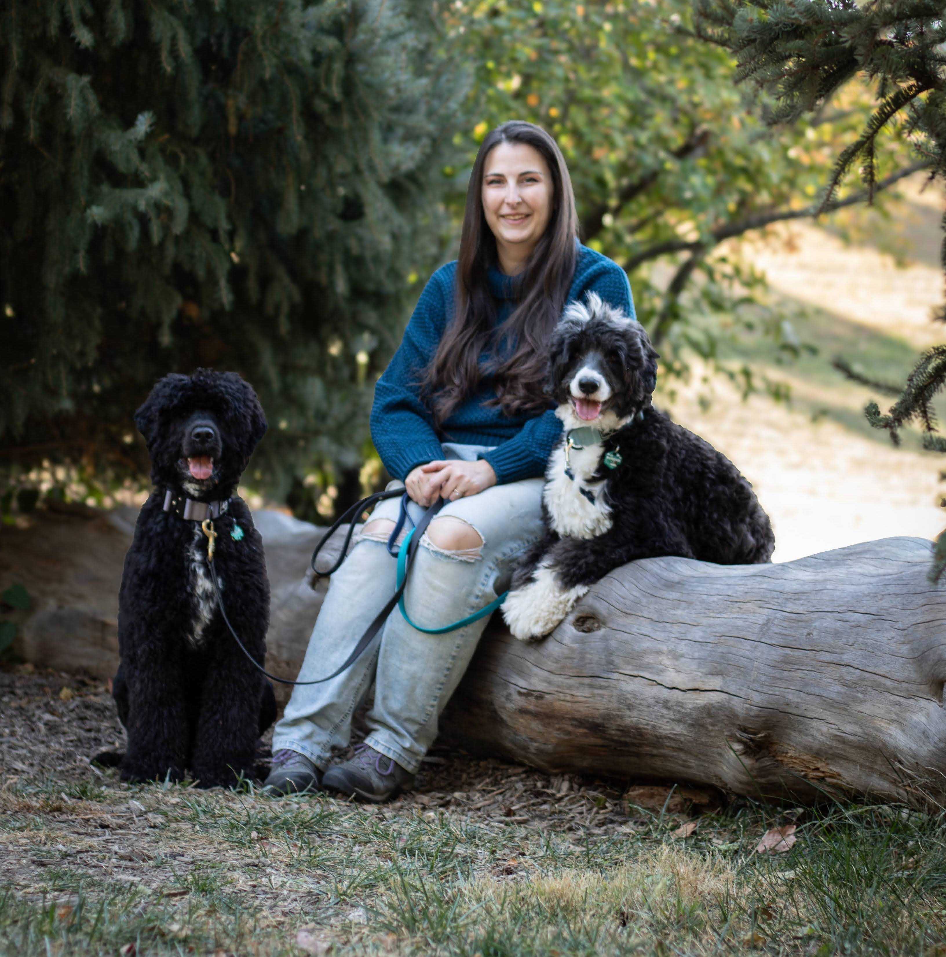 A woman with long dark hair wearing a darker teal sweater and jeans smiling at the camera with two Portuguese Water Dogs on either side of her. On her left, a fluffy black dog is sitting, and on her right, a similar black and white dog is partly lying on the log, also looking at the camera.