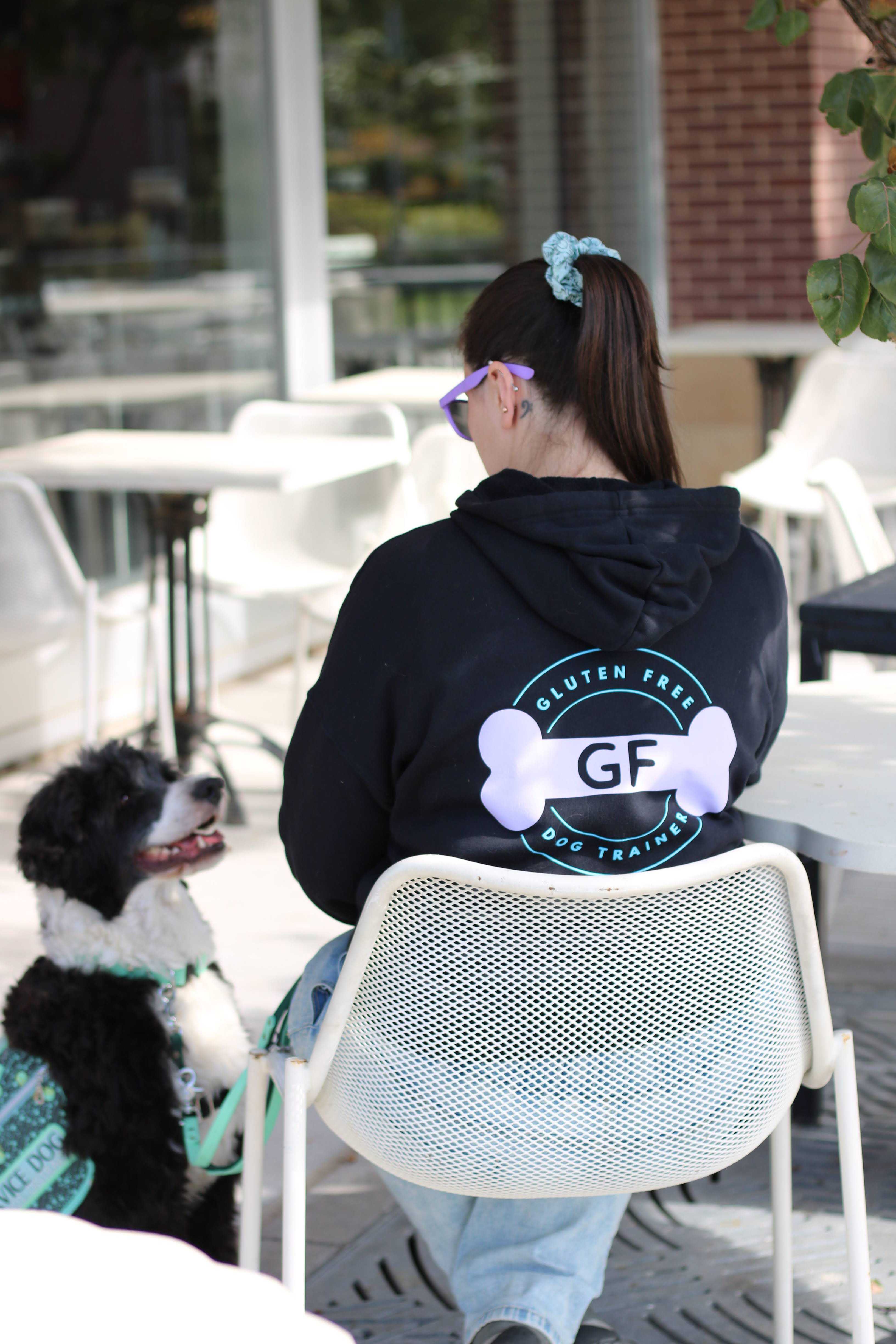 A woman wearing a black hoodie with the Gluten Free Dog Trainer logo on the back sits at an outdoor café table. Her hair is tied back with a blue scrunchie, and she wears purple sunglasses. A black and white Portuguese Water Dog wearing a seafoam green service dog vest looks up at her attentively.