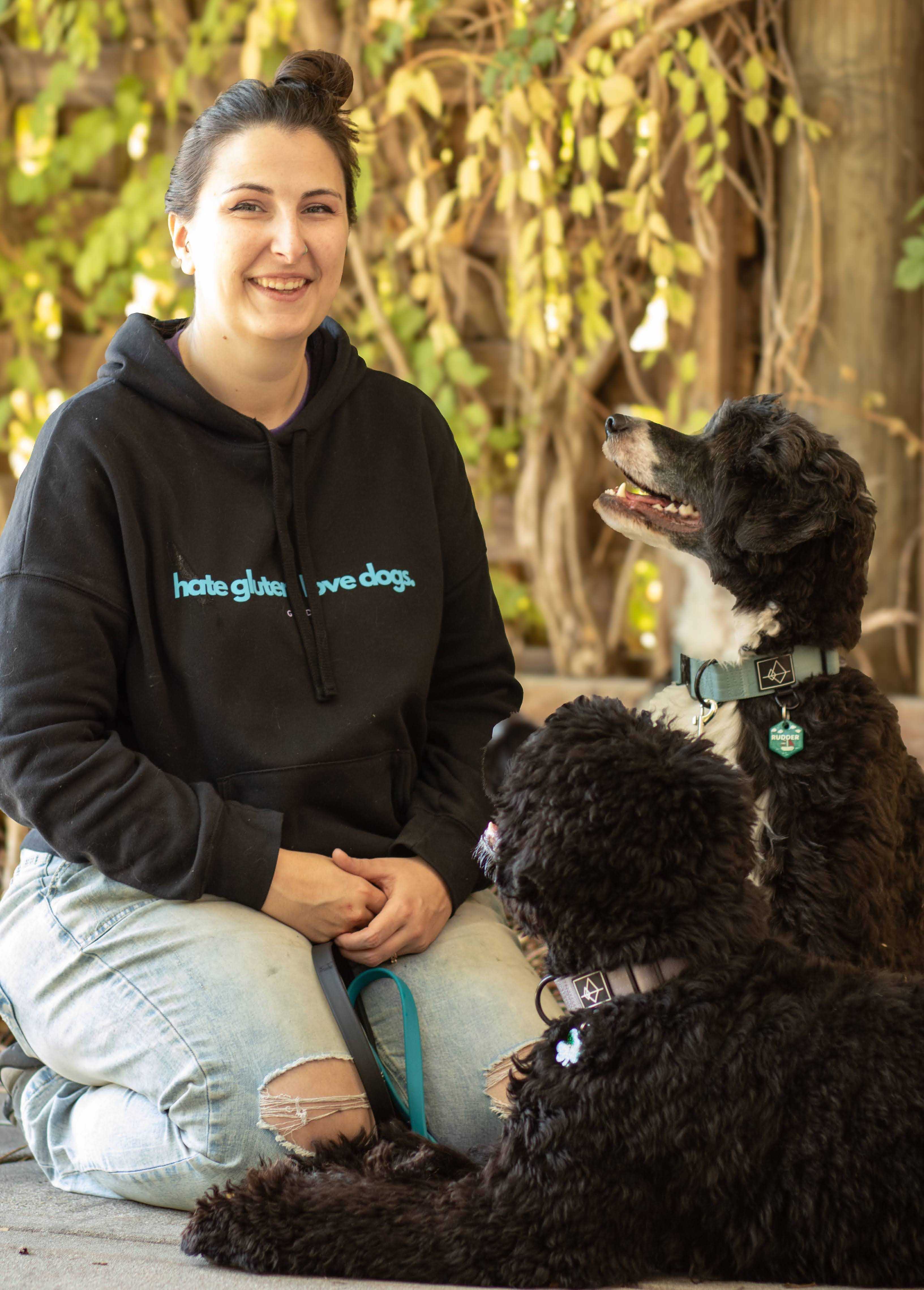 A woman smiling seated on the ground in an outdoor setting, surrounded by two Portuguese Water Dogs. The woman is wearing a black hoodie with blue text stating "hate gluten. love dogs." and ripped jeans. They have their hair tied up with a clip. One dog is sitting and looking up at the woman, while the other is lying down and facing her. The scene is set against a backdrop of green and yellow leaves with some tree branches visible.